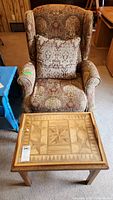 View of brown tapestry wingback chair with matching ornamental pillow and removable wooden tray table with geometric wood inlay pattern on top.