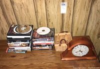 Photo showing mantel clock, basket, and stacks of DVDs and CDs on wood surface with wood-paneled background.