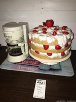 Photo showing the Mr. Coffee coffee maker next to the ceramic strawberry cake dish on a Coca Cola glass cutting board, all arranged together on a flat surface.
