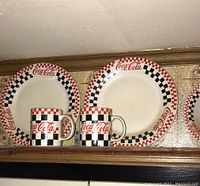 Two Coca-Cola ceramic plates and two mugs with red and black checkered design on the rims and bodies.