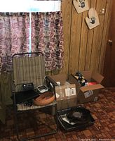 Chair with assorted items including cooking pan, skillet, blood pressure monitor, iron, boxes with electronics or small appliances, and two plant pot bottoms below window with Coca Cola curtains