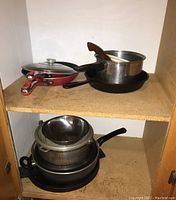 Cookware including red frying pan, stainless pot with lid, cast iron skillets, and metal strainer bowl on wood shelves.