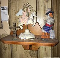 Small wooden wall shelf holding an angel figurine riding a white carousel horse, rustic carved cross, duck figurine dressed in fabric clothes and hat, and a small metallic keychain with keys.