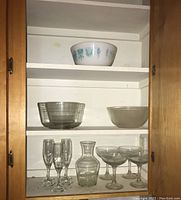 Glassware and bowls displayed in cabinet: five bowls (one milk glass with blue-green folk art design, one ribbed glass, and three clear glass bowls), one clear glass carafe, five champagne flutes, and four margarita glasses.
