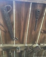 Image shows multiple rusty iron farm tools hanging on a wooden fence background including hooks, latches, and bolts as decorative farm implement items.