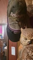 Photo showing three men's hats stacked vertically on a wall-mounted rack, with the Remington camo hat on top, and the owl statue figurine in front. The background includes a wooden wall and a glimpse of a framed print.