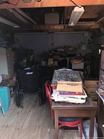 Full room view showing wheelchair, file cabinets, wooden desk with papers, boxes, and assorted storage items