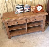 Front view of wooden media shelf with two drawers and open shelves, showing some DVDs and a clock on top for scale.