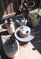 Gas cans, watering cans, and large metal fryer pot on concrete floor near wooden fence.