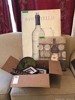 Two wine-themed wall art pieces positioned behind a box on a sofa; box contains additional wine decor items.