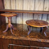 Two round wood display tables on a wooden surface against a paneled wall, showing different designs and sizes.