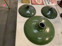 Three green antique metal pool hall shades displayed on a white surface; one larger shade measuring 17.75 inches diameter and two smaller 12 inch diameter shades, showing rust and wear.