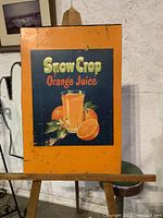 Front view of antique metal sign with Snow Crop Orange Juice advertisement showing orange border and juice glass illustration with fruit.