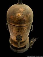 Front angled view of large hammered copper samovar lamp showing spout and age patina on copper surface with attached lamp socket and wooden base visible.