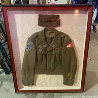 Framed display showing field jacket mounted below garrison cap behind glass in wood frame