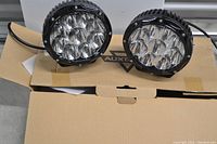 Pair of round LED vehicle lights placed on top of a cardboard box showing front lenses and housing.