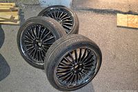 Three black multi-spoke rims with tires laid on pavement, showing overall set and condition