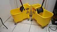 Two large yellow Rubbermaid mop buckets with mop wringers and caster wheels placed side by side on tiled floor, with caution wet floor symbol visible on the buckets.