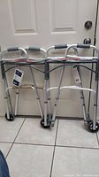 Front view of two new Drive walkers with tags standing side by side showing hand grips, wheels and locking mechanisms.