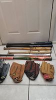 Photo of 5 baseball bats and 8 baseball gloves arranged on floor in front of a white door. Bats include black and natural wood Louisville Sluggers and a green/orange alloy bat. Gloves in various shades of brown leather.