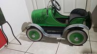 Front and side view of green roadster pedal car showing overall body, wheels, and missing front left silver hubcap.