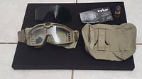 Wide shot showing ESS Influx tan goggles with clear lens, extra dark lens, carrying case, lens cleaner, and information insert arranged on black surface.