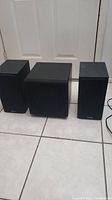 One subwoofer placed between two bookshelf speakers on tiled floor against a closed white door, showing overall condition and relative size.