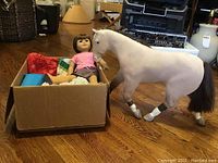 American Girl doll with light brown hair and bangs, seated in a cardboard box, with the white horse figure standing nearby on the floor.