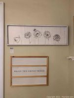 Photo showing both wood art pieces mounted on a wall. The top piece is a rectangular panel with black line art flowers. The bottom piece is a smaller framed quotation piece.