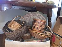 Photo of multiple assorted vintage wicker baskets stacked inside a box under a table, showing different sizes and weaving styles in natural brown tones.