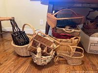 Multiple varied picnic and garden baskets in woven, wood, and coated metal materials arranged on floor near wooden table and chairs