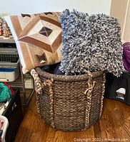 Photo showing two pillows (brown and cream geometric with cow fur side, gray shaggy) sitting in large woven seagrass basket with black wash finish and broken handles