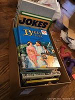 View of a box full of books including The Bible Story volume II with colorful illustrated cover, a jokes book, and other books stacked inside a cardboard box, floor setting.