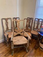 Set of six wooden dining chairs with natural rush woven seats, two captain chairs with armrests and four side chairs without armrests arranged in a corner against cream walls and hardwood floor.