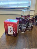 Photo of six Coca-Cola branded drinking glasses with purple magenta stained glass design and a large matching glass pitcher alongside the original packaging box showing a Rockwell commemorative Coca-Cola tankard image