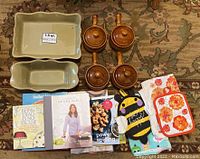 Set including stoneware bakeware, crock bowls with lids, oven mitts, dish towels, and cookbooks arranged on carpet.