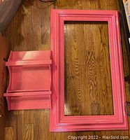 Top-down view of pink painted wooden wall shelf and large frame on hardwood floor