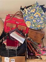 Photo shows assorted handbags stacked including large red Vera Bradley floral drawstring backpack, red canvas and tan leather buckle bag, woven straw handbag, and various small bags.