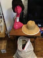 Pink mannequin head with brown faux fur Hiki by New Wadley hat on wooden table next to natural straw hat. White plastic bag below containing additional hats including leopard print.