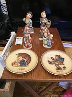 All six Hummel figurines displayed on wooden surface with two decorative plates in front. Plates show dates 1973 and 1982 and typical Hummel artwork.