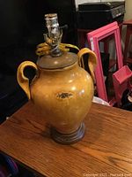 Full view of mustard yellow ceramic table lamp with metal top and exposed lamp socket, placed on wooden surface in a room with various objects in background.