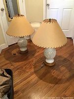 Pair of large white ceramic lamps with beige stitched shades, shown whole resting on wooden floor