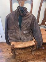 Front view of brown leather jacket resting on a wooden chair, showing zipper front, collar, chest zipper pocket, and two flap pockets.