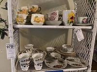Two shelves of plates and cups with floral, rustic, and traditional railroad patterns, displayed on white wicker shelves.