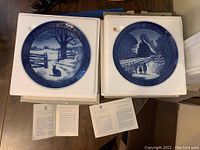 Two Royal Copenhagen Christmas plates placed on white padded holders alongside information cards, showing front of plates with winter designs and dates 1971 and 1973