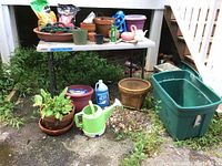Wide view showing clay and plastic pots, expandable green hose, various gardening materials on table, large green plastic bin, woven basket planter, and bottle of windshield wiper fluid on ground.