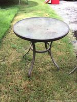 Front angle view of round glass topped metal patio table on grass, showing integral glass surface and metal legs.