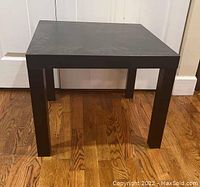 Black square IKEA particle board table standing on hardwood floor, front angle showing all four legs and top surface.