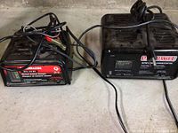 Two black Motomaster battery chargers on a concrete floor. One charger shows a red label (manual 6/2A 6 & 12V), the other a black label with charge rate gauge (10A / 2A). Cables and clamps visible. Shows wear and dirt on surfaces.