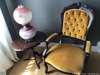 View of antique upholstered armchair with yellow velvet tufted backrest next to small hardwood dropleaf table and decorative lamp